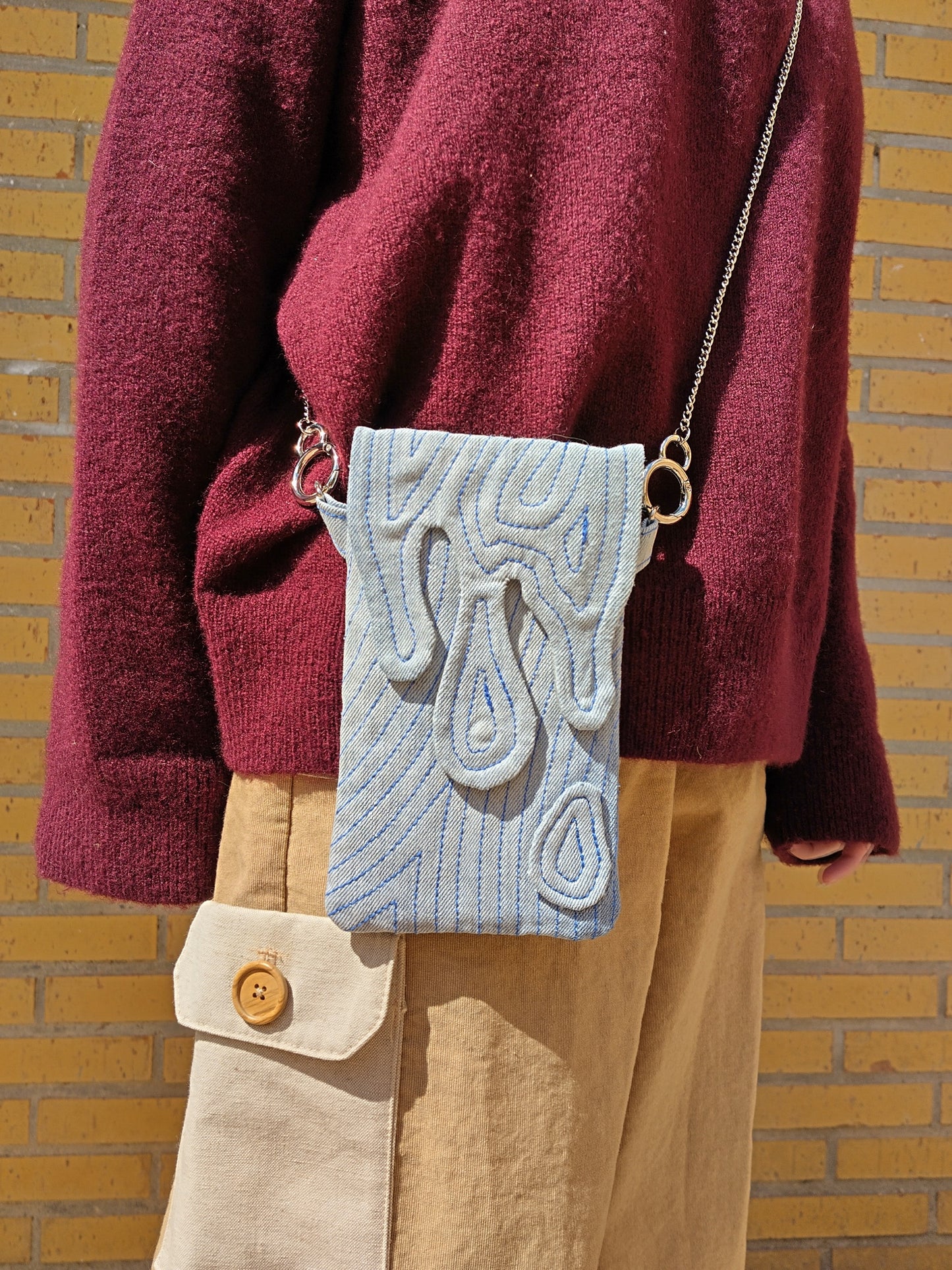 Bolso rectangular colgable, de tejido vaquero con cierre en forma de gotas cayendo. Con cadena desmontable de metal plateada.