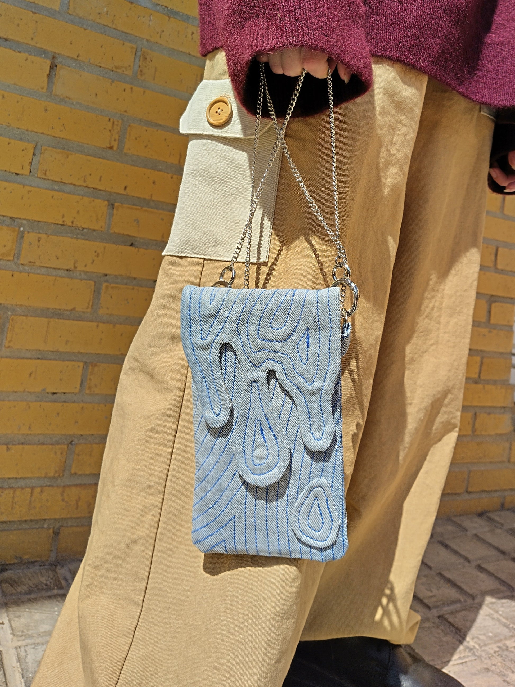 Bolso rectangular colgable, de tejido vaquero con cierre en forma de gotas cayendo. Con cadena desmontable de metal plateada.