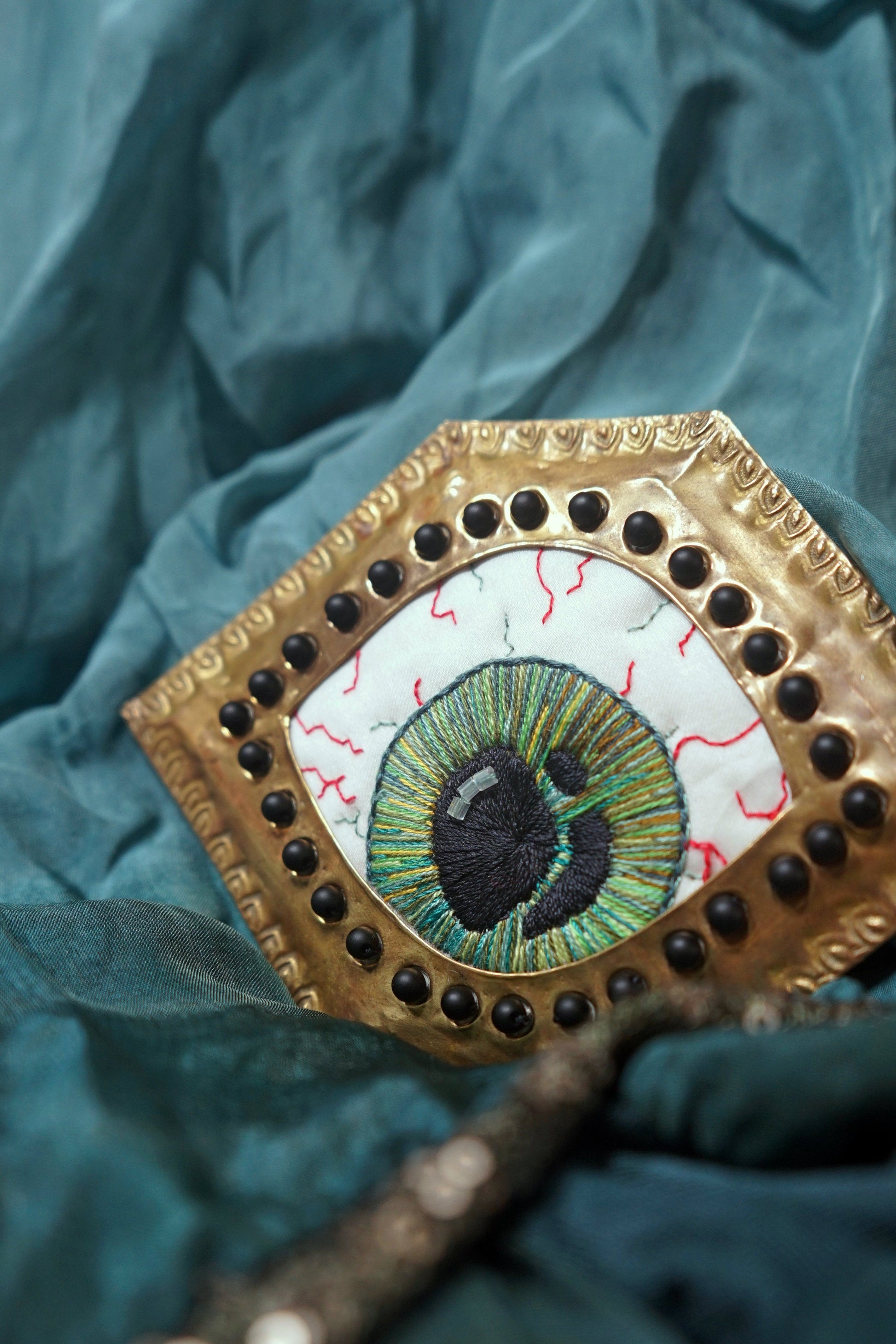 Cuadro de ojo verde bordado en marco dorado geométrico ornamentado con piedras negras.