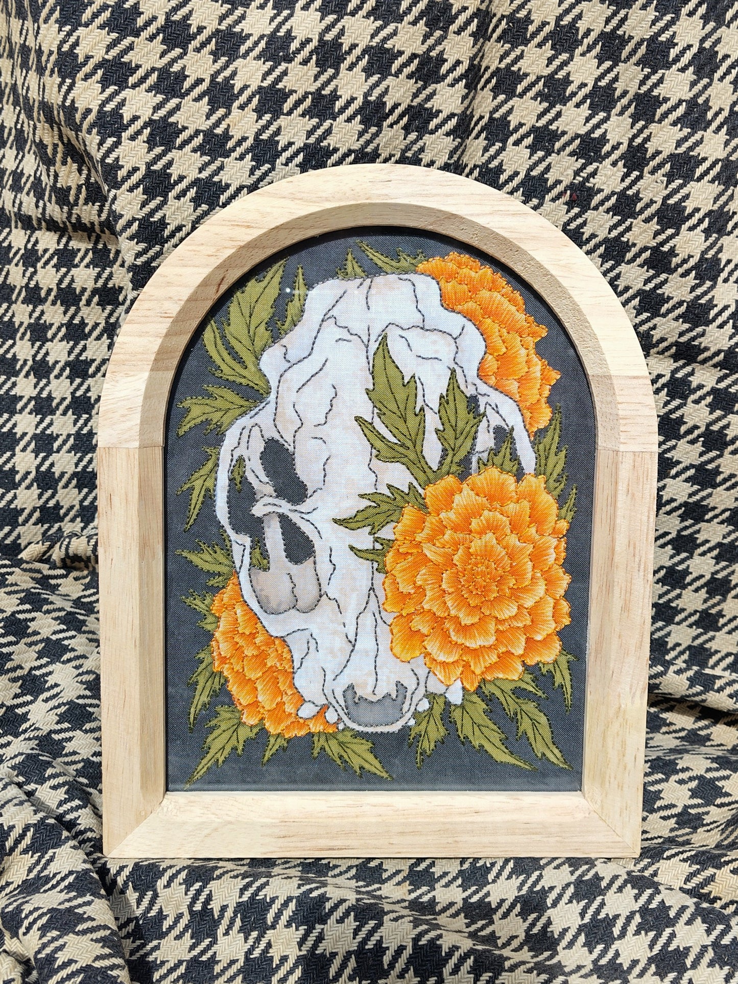 Cuadro pintado y bordado de calavera de gato anatómica rodeada de tagetes naranjas sobre fondo negro.