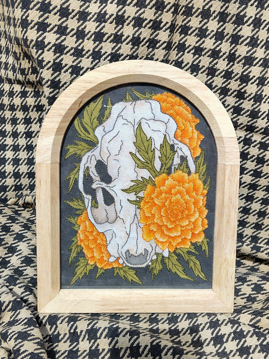 Cuadro pintado y bordado de calavera de gato anatómica rodeada de tagetes naranjas sobre fondo negro.